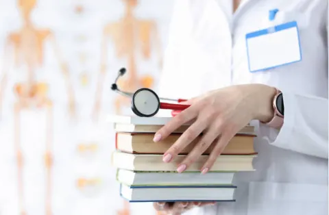 A clinician in a white coat holding a stack of books and a stethoscope